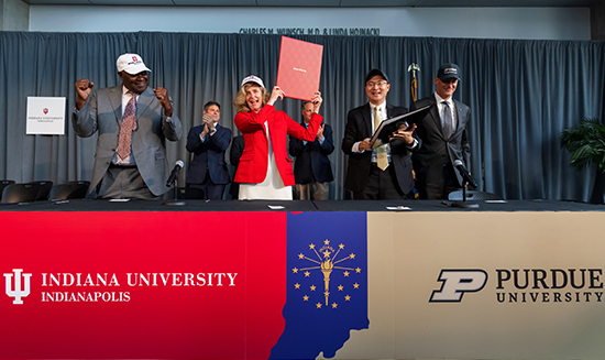 Presidents and trustees of IU and Purdue stand behind a draped table clapping at the transition event.